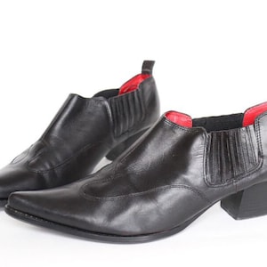 Puede incluir: Un par de botines de cuero negro con punta puntiaguda y un pequeño tacón. Las botas tienen un forro rojo y un estilo de poner y quitar.