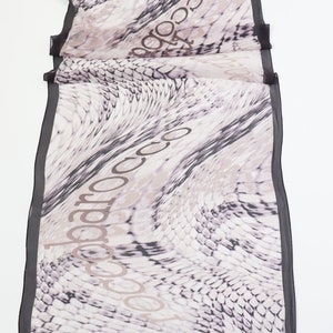 May include: A white and black patterned scarf with a black border. The scarf has the word "tocca" printed on it in a brown colour.