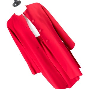 Könnte beinhalten: Ein leuchtend roter Anzug, bestehend aus einem langen Mantel und einer passenden Hose, auf einer Schaufensterpuppe präsentiert. Der Mantel hat eine Knopfleiste und lange Ärmel. Das Outfit ist einfarbig und wirkt elegant und raffiniert.