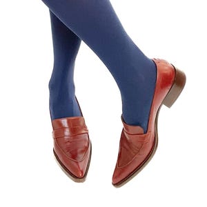 Puede incluir: Un par de mocasines de cuero rojo con puntera puntiaguda y un pequeño tacón. Los mocasines se llevan con medias azul oscuro.