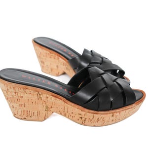 Puede incluir: Sandalias de cuña de cuero negro con un diseño de correas trenzadas. Las sandalias tienen una plataforma y un tacón de cuña de corcho. El nombre de la marca está impreso en la plantilla. La altura del tacón es de aproximadamente 7,6 cm.