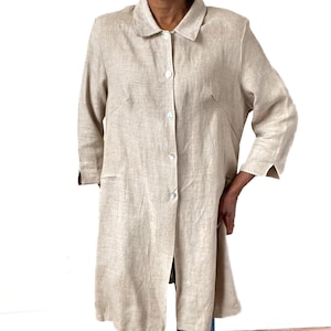 May include: A beige linen coat with a collared neckline and three-quarter sleeves. The coat has a button-down front and two front pockets. The knee-length garment is made of a lightweight fabric, suitable for layering, and is a neutral colour.