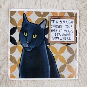 Könnte beinhalten: Eine schwarze Katze mit grünen Augen sitzt auf einem braunen und gelben gemusterten Hintergrund. Ein Schild über der Katze lautet "Wenn eine schwarze Katze deinen Weg kreuzt, bedeutet das, dass sie irgendwohin geht."