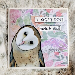 May include: A white and brown barn owl with large, dark eyes is depicted on a square ceramic tile. The owl is looking to the right. The background is a watercolor floral pattern in shades of pink, purple, green, and orange. The tile has the text "I REALLY DON'T GIVE A HOOT" printed on it in black.
