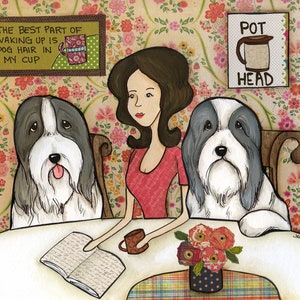 Könnte beinhalten: Eine Frau sitzt an einem Tisch mit zwei Bearded Collie Hunden. Die Frau liest ein Buch und hält eine Tasse. Die Hunde schauen die Frau an. Der Tisch hat eine Blumendecke und eine Vase mit Blumen. Die Wand hinter dem Tisch hat eine Blumentapete. Der Text an der Wand lautet "The best part of waking up is dog hair in my cup".