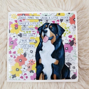 May include: A white ceramic tile with a floral pattern background. A Bernese Mountain Dog is sitting in the center of the tile. The dog is looking at the camera with its tongue sticking out. The tile has text that reads: "DOGS MAKE YOU LAUGH A LITTLE LOUDER SMILE A LITTLE BRIGHTER AND LIVE YOUR BEST LIFE EVER".
