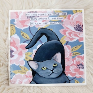 May include: A blue and gray cat with green eyes looks at the camera. The cat is sitting on a floral background with pink and yellow flowers. The text "NOTHING MAKES YOU SMILE MORE THAN LOOKING AT THE FACE OF A CAT THAT LOVES YOU" is written in black on a white background.