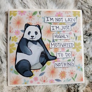 May include: Square ceramic tile with a panda bear illustration. The panda is black and white, sitting with its legs extended. The tile has a floral background and text that reads "I'm not lazy, I'm just highly motivated to do nothing."