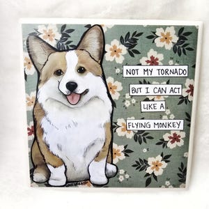 May include: Square ceramic tile featuring a smiling corgi illustration with brown, white, and tan fur. The background is a floral pattern in shades of green, cream, and brown. Text on the tile reads: "NOT MY TORNADO BUT I CAN ACT LIKE A FLYING MONKEY."
