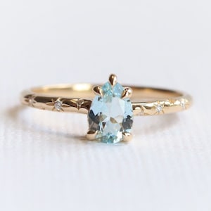May include: A gold ring featuring a pear-shaped, light blue gemstone. The band is adorned with star and dot patterns and small accent stones. The ring is set against a white background, highlighting its delicate design.