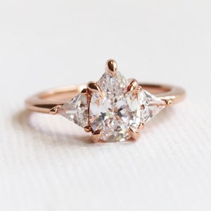 Puede incluir: Un anillo de compromiso de oro rosa con una piedra central grande de forma ovalada y dos piedras triangulares m&aacute;s peque&ntilde;as a cada lado.