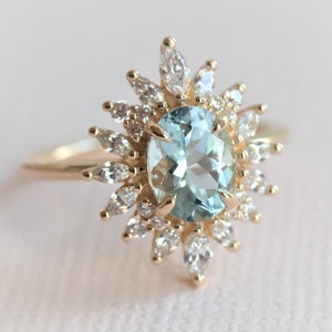 May include: A gold ring featuring an oval, light blue gemstone surrounded by smaller, clear gemstones. The ring's design resembles a sunburst, with the central stone as the focal point. The band is a simple, polished gold.