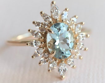 May include: A gold ring featuring an oval, light blue gemstone surrounded by smaller, clear gemstones. The ring's design resembles a sunburst, with the central stone as the focal point. The band is a simple, polished gold.