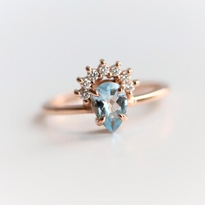 May include: A delicate rose gold ring with a pear-shaped aquamarine gemstone surrounded by a halo of small diamonds.