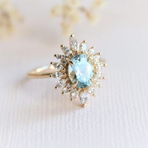 May include: A gold ring featuring a light blue oval gemstone surrounded by smaller white gemstones in a sunburst design. The ring has a delicate band and is set against a soft, blurred background.