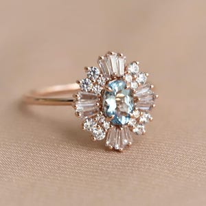 May include: Rose gold ring featuring an oval, light blue gemstone surrounded by a halo of smaller white and baguette-cut stones. The ring's design is reminiscent of a floral burst, with a delicate band.