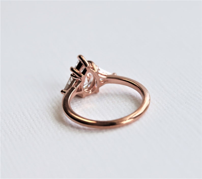 Puede incluir: Un anillo de compromiso de oro rosa con una piedra central grande de forma ovalada y dos piedras m&aacute;s peque&ntilde;as a cada lado. El anillo est&aacute; engastado en un dise&ntilde;o delicado e intrincado.