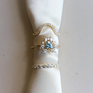 May include: Three gold rings with clear and blue gemstones. The rings are stacked on top of each other on a white fabric background.