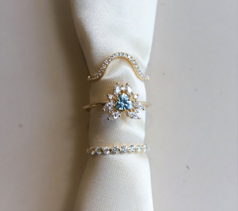 May include: Three gold rings with clear and blue gemstones. The rings are stacked on top of each other on a white fabric background.