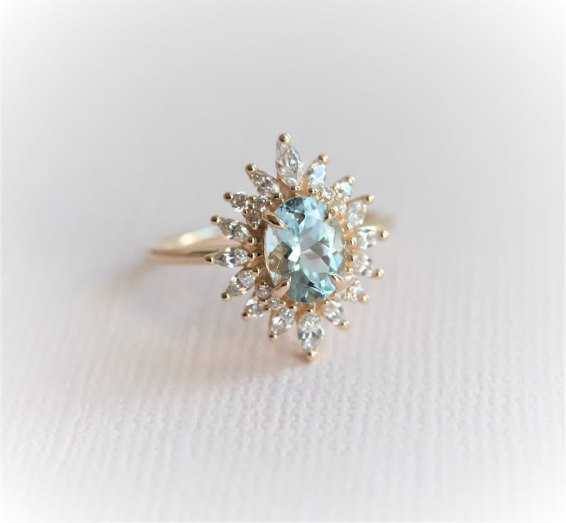May include: A gold ring featuring an oval, light blue gemstone surrounded by smaller, clear gemstones. The ring's design resembles a sunburst, with the central stone as the focal point. The band is a simple, polished gold.