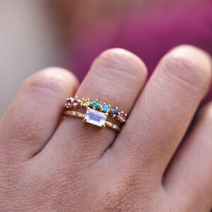 May include: A gold ring with a rectangular clear gemstone and a smaller band of colorful gemstones. The ring is on a person's finger.