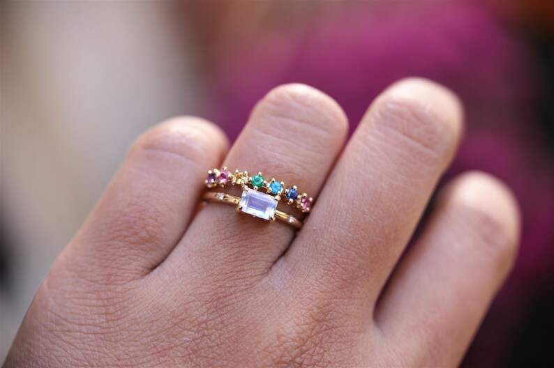 May include: A gold ring with a rectangular clear gemstone and a smaller band of colorful gemstones. The ring is on a person's finger.