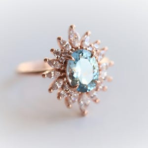 May include: A rose gold ring featuring an oval, light blue gemstone surrounded by smaller, clear gemstones. The ring's design resembles a sunburst, with the central stone as the focal point. The band is a simple, polished rose gold.