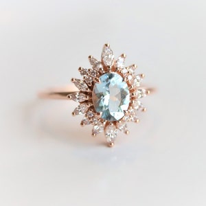 May include: A rose gold ring featuring an oval, light blue gemstone at the center, surrounded by a halo of smaller, marquise-cut and round white gemstones. The ring's band is a delicate rose gold.