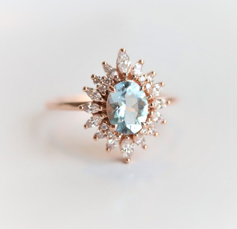 May include: A rose gold ring featuring an oval, light blue gemstone at the center, surrounded by a halo of smaller, marquise-cut and round white gemstones. The ring's band is a delicate rose gold.