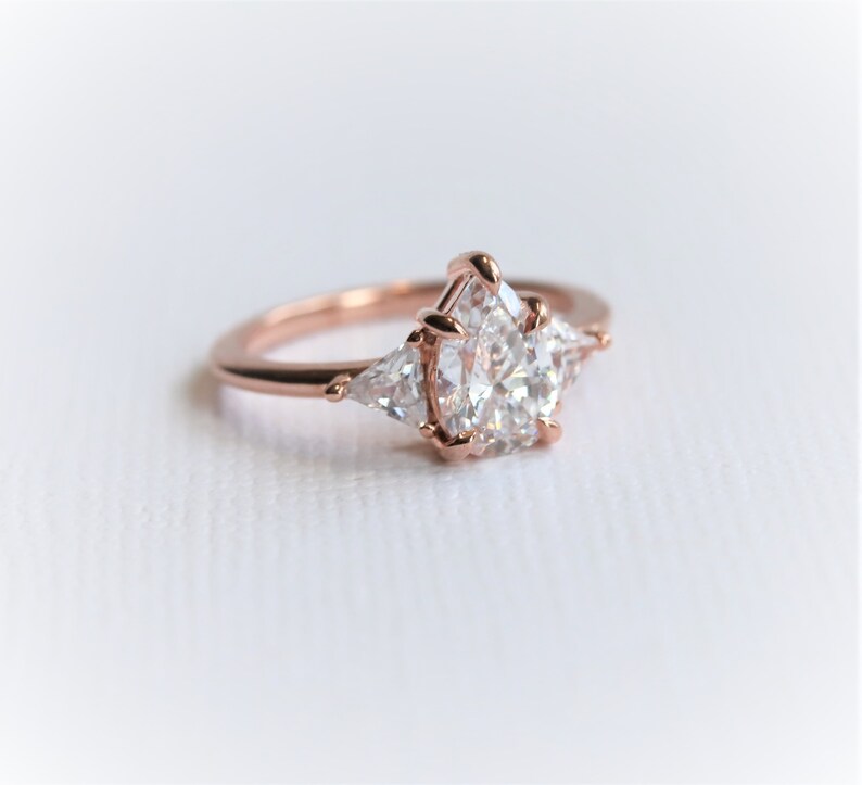 Puede incluir: Un anillo de compromiso de oro rosa con una piedra central en forma de pera y dos piedras de acento triangulares m&aacute;s peque&ntilde;as.