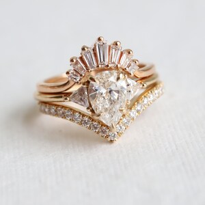 Puede incluir: Un anillo de compromiso de oro con una piedra central de diamante en forma de pera, rodeado de diamantes m&aacute;s peque&ntilde;os en una banda delicada en forma de V. El anillo est&aacute; acentuado por un halo de diamantes de corte baguette.