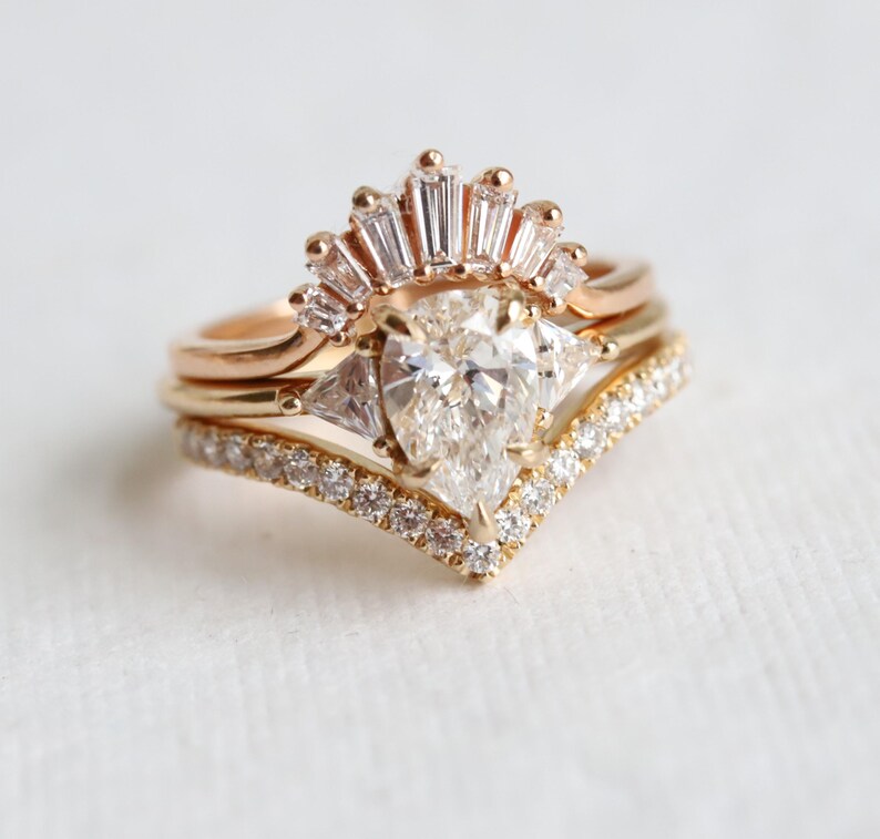 Puede incluir: Un anillo de compromiso de oro con una piedra central de diamante en forma de pera, rodeado de diamantes m&aacute;s peque&ntilde;os en una banda delicada en forma de V. El anillo est&aacute; acentuado por un halo de diamantes de corte baguette.