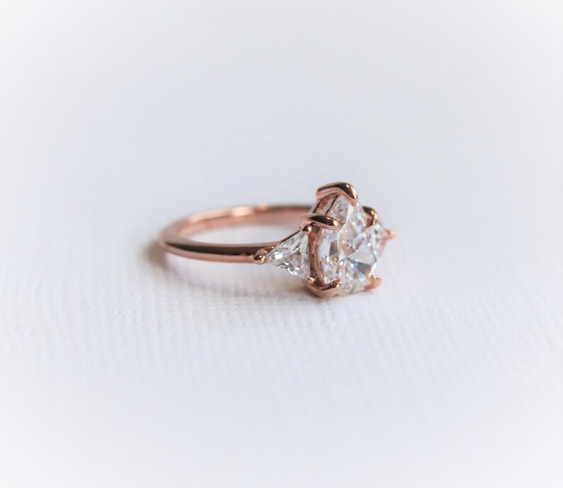 Puede incluir: Un anillo de compromiso de oro rosa con una piedra central grande, redonda y transparente y dos piedras triangulares m&aacute;s peque&ntilde;as y transparentes a cada lado.