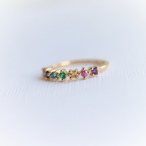May include: A gold band ring with a row of colorful gemstones. The gemstones are set in a prong setting and are arranged in a rainbow pattern. The ring is a delicate and feminine piece of jewelry.