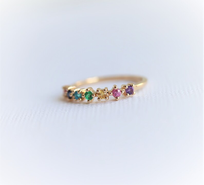 May include: A gold band ring with a row of colorful gemstones. The gemstones are set in a prong setting and are arranged in a rainbow pattern. The ring is a delicate and feminine piece of jewelry.