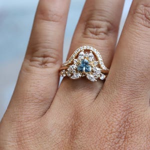 May include: A gold ring with a blue gemstone surrounded by smaller white gemstones. The ring is set on a finger.