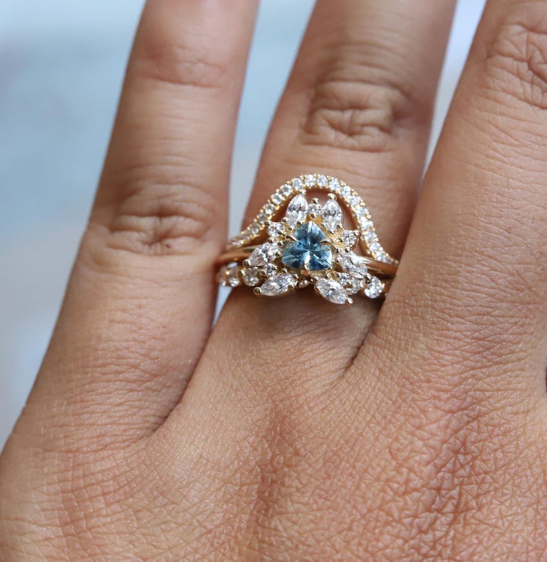 May include: A gold ring with a blue gemstone surrounded by smaller white gemstones. The ring is set on a finger.