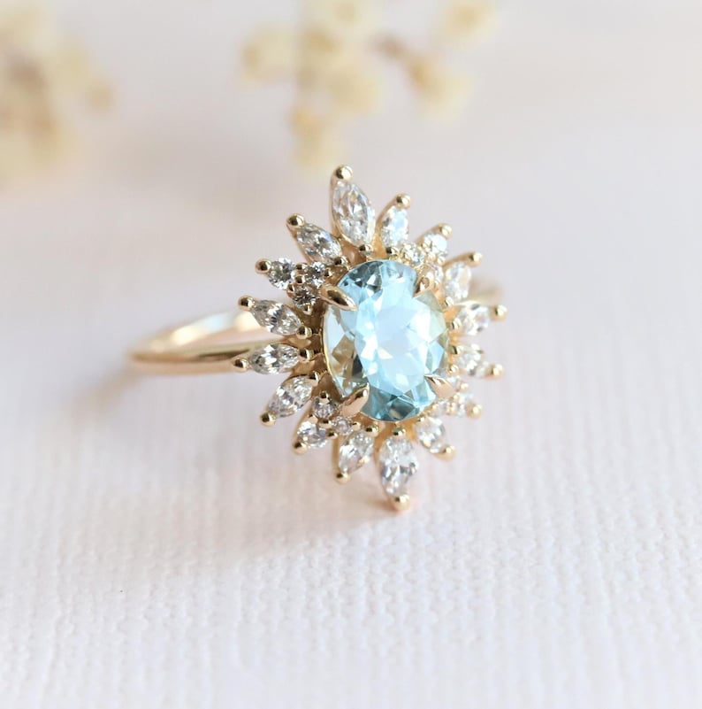 May include: A gold ring featuring a light blue oval gemstone surrounded by smaller white gemstones in a sunburst design. The ring has a delicate band and is set against a soft, blurred background.