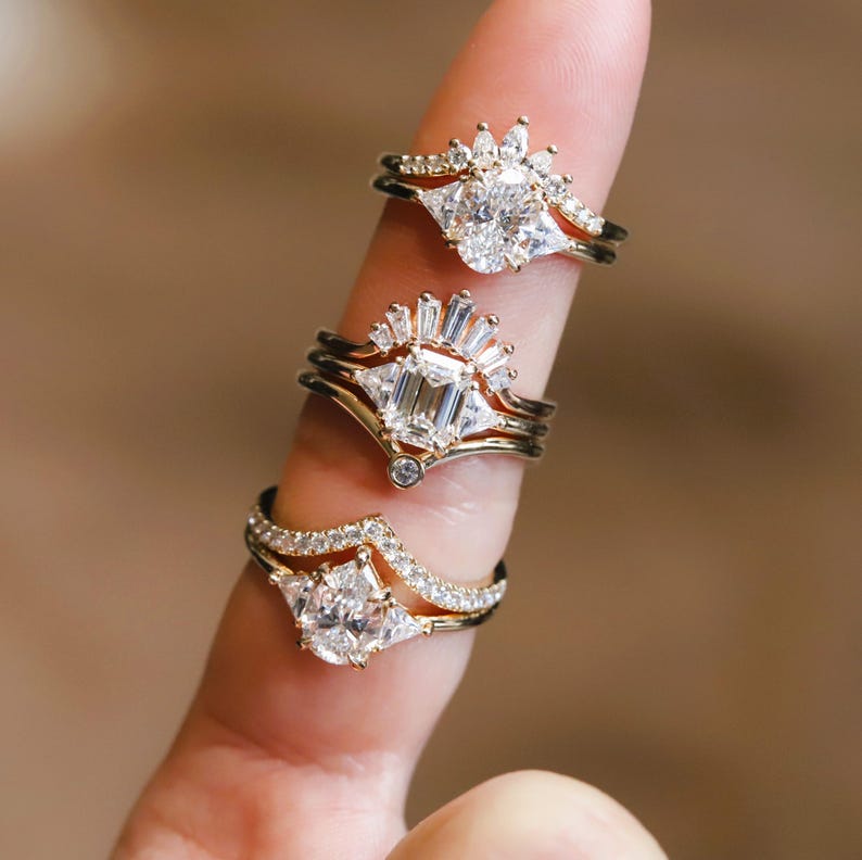 Puede incluir: Tres anillos de oro con diferentes engastes de diamantes. El anillo superior presenta un gran diamante ovalado rodeado de diamantes m&aacute;s peque&ntilde;os. El anillo del medio presenta un gran diamante de talla esmeralda rodeado de diamantes baguette m&aacute;s peque&ntilde;os. El anillo inferior presenta un gran diamante en forma de pera rodeado de diamantes triangulares m&aacute;s peque&ntilde;os.
