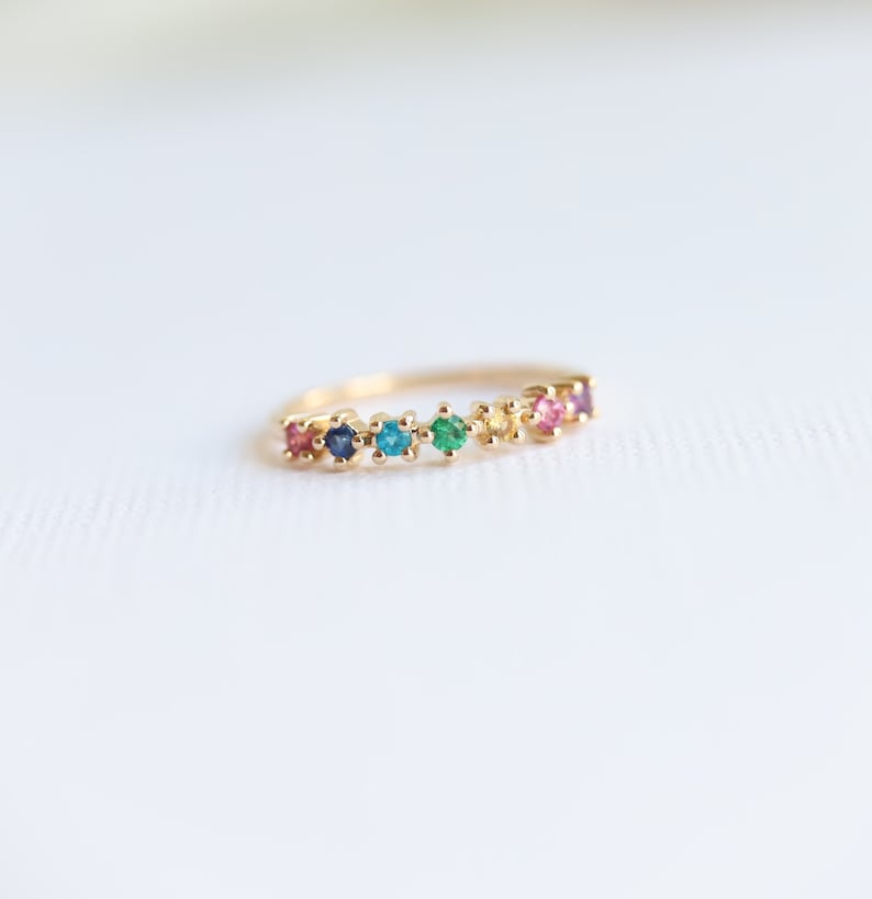 May include: A gold band ring with a rainbow of gemstones. The ring features a row of small, colorful stones set in a gold band.
