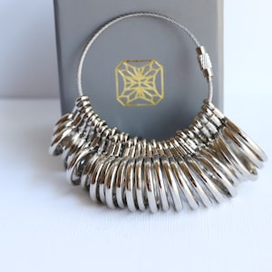 May include: A silver ring sizer with a chain and a clasp. The sizer has a variety of ring sizes to help determine the correct fit.