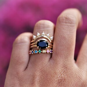 May include: A set of three gold rings with a black oval gemstone in the center ring. The top ring has a row of small white gemstones. The bottom ring has a row of colorful gemstones.