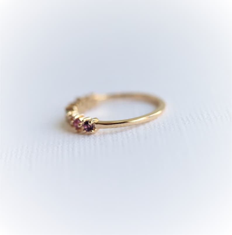 May include: A delicate gold band ring with a row of small, colorful gemstones. The ring is set against a white background.