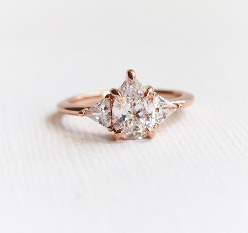 Puede incluir: Un anillo de compromiso de oro rosa con una piedra central grande de forma ovalada y dos piedras triangulares m&aacute;s peque&ntilde;as a cada lado.