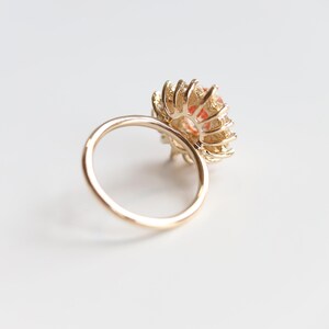 May include: A gold ring with a large, round, orange gemstone center. The gemstone is surrounded by a halo of smaller, gold-colored elements, creating a flower-like design. The ring band is a simple, polished gold.