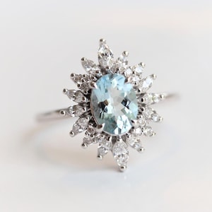May include: A silver ring featuring a light blue oval gemstone surrounded by a halo of smaller, marquise-cut white gemstones. The ring's design resembles a sunburst.