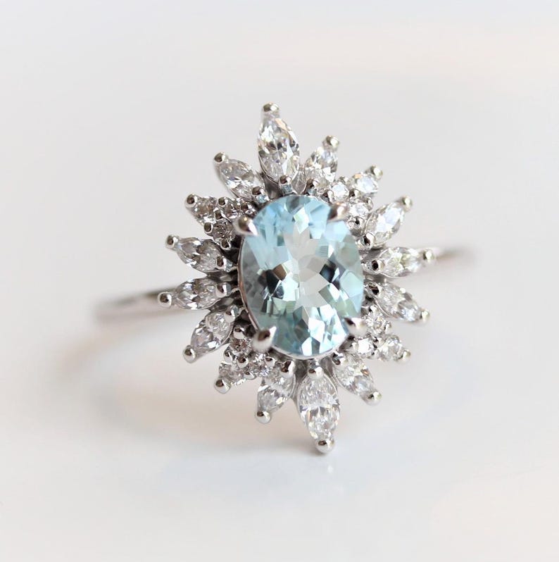 May include: A silver ring featuring a light blue oval gemstone surrounded by a halo of smaller, marquise-cut white gemstones. The ring's design resembles a sunburst.