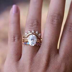 May include: A set of three gold rings with a large oval-shaped center stone. The center ring has a row of small pearls on the top. The bottom ring has a row of colorful gemstones.