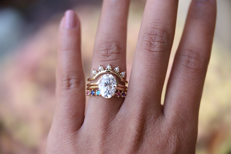 May include: A set of three gold rings with a large oval-shaped center stone. The center ring has a row of small pearls on the top. The bottom ring has a row of colorful gemstones.