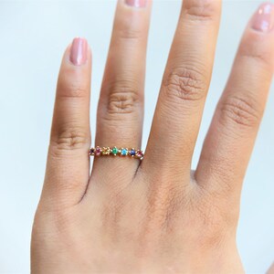 May include: A gold band ring with a row of colorful gemstones. The ring is on a person's finger.
